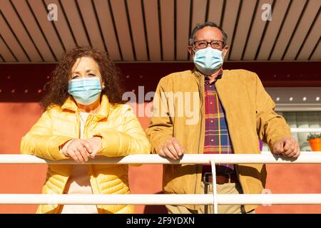 Porträt von zwei älteren Bürgern, die von der Veranda ihres Hauses aus das Äußere beobachten. Sie tragen eine Schutzmaske, um das Covid-19-Virus zu verhindern. Stockfoto