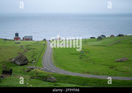 Straße, die zum faroesischen Dorf Trollanes führt, von einem Hügel in der Nähe betrachtet. Grüne Hügel, trübes, nebliges Wetter im wilden Norden. Eher ruhiges Meer und Ty Stockfoto