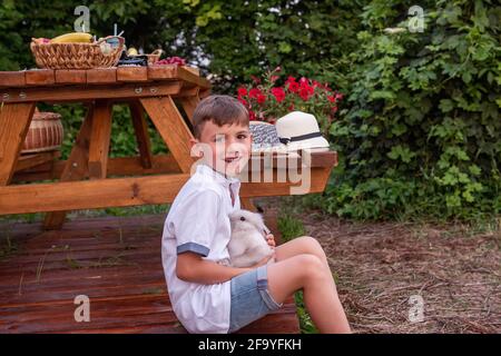 Das Porträt eines kleinen Jungen in einem Strohhut sitzt zu Hause in der Nähe eines LKW-Anhängers auf einem Holzboden und hält ein flauschiges Kaninchen in den Händen. Lacht, spielt mit Stockfoto