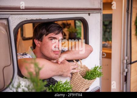 Das Porträt eines jungen Mannes blickt aus dem Fenster eines Wohnwagens mit Anhänger zwischen grünen Pflanzen. Die Brünette ruht mit seiner Familie außerhalb der Stadt auf t Stockfoto