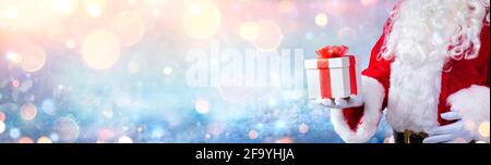 Weihnachtsmann hält Ein Weihnachtsgeschenk mit verschneiten Hintergrund und Golden Lights Stockfoto
