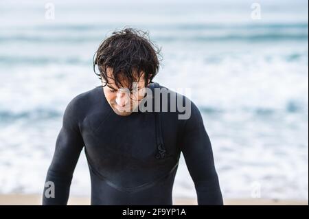 Mittelgroßer, nasser Mann mit Tauchanzug, der aus dem Meer herauskommt und nach unten blickt. Stockfoto