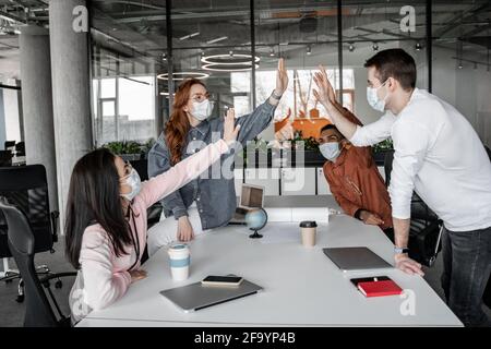 Interracial Studenten in medizinischen Masken geben hohe fünf in der Nähe Gadgets Auf dem Schreibtisch Stockfoto