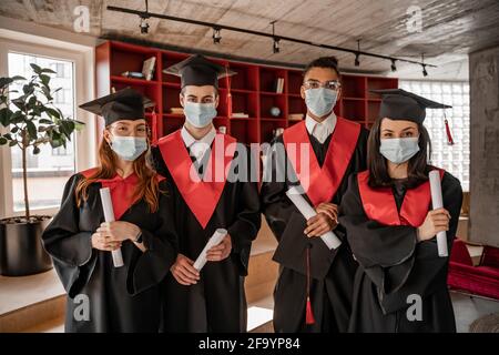 Interracial Studenten in medizinischen Masken, Abschlusskleider und Kappen mit Diplom, Senior 2021 Stockfoto