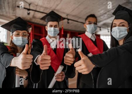 Interracial Studenten in medizinischen Masken, Kleidern und Kappen mit Diplom, während sie Daumen nach oben zeigen, Abschlussklasse 2021 Stockfoto