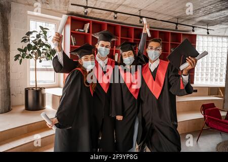 Multiethnische Studenten in medizinischen Masken, Kleidern und Kappen mit Diplom, Abschlussklasse 2021 Stockfoto