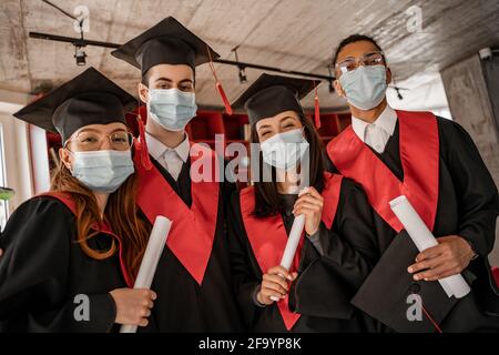 Multiethnische Studenten in medizinischen Masken, Abschlusskleidern und Caps mit Diplom, Senior 2021 Stockfoto