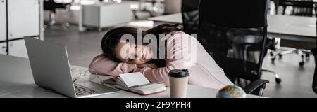 Ein müder Student schläft auf dem Schreibtisch neben dem Laptop und dem Papierbecher und dem Banner Stockfoto