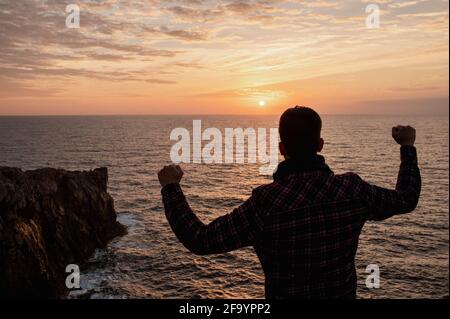 Rückansicht Silhouette eines unverkennbaren Mannes auf der Spitze einer felsigen Klippe, der gegen das Meer steht und den Sonnenuntergang betrachtet und die Arme mit der Siegesfaust hebt. Stockfoto