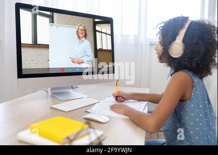 Homeschooling und e-studing Konzept. Nettes biracial Mädchen beobachten Online-Kurse, Webinare, Online-Lernen mit einem Senior-Lehrer auf dem Bildschirm. Schulkind, das auf der Strecke studiert Stockfoto