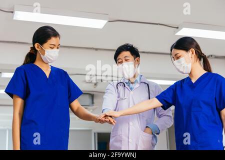 Gruppe von asiatischen männlichen und weiblichen Beschäftigten im Gesundheitswesen wie Arzt Und Krankenschwester trägt OP-Maske und Uniform mit Stethoskop stehend Beim Stapeln der Hände im Krankenhaus Stockfoto