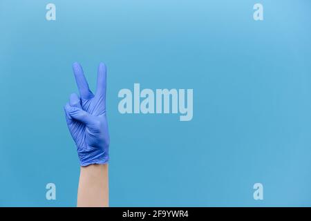 Nahaufnahme der männlichen Hand in medizinischen Schutzhandschuhen zeigt zwei Finger nach oben, isoliert in blauen Studio-Hintergrund mit Kopieplatz, drückt Feier Stockfoto