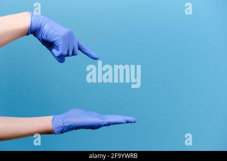 Nicht erkennbarer junger Mann, der sich öffnet und auf die Handfläche zeigt, trägt medizinische Schutzhandschuhe, isoliert auf der blauen Wand im Hintergrund des Studios Stockfoto