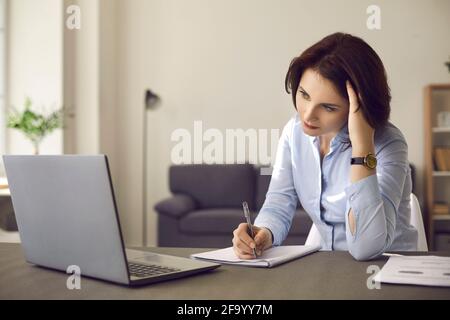 Frau, die an einem Schreibtisch sitzt und einen Laptop vor sich hat und ernsthaft in einem Notebook schreibt. Stockfoto