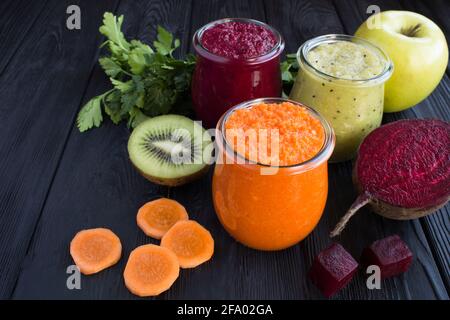 Verschiedene Gemüse- und Obstsmoothies oder Püree in den kleinen Gläsern auf dem schwarzen Holzhintergrund. Nahaufnahme. Stockfoto