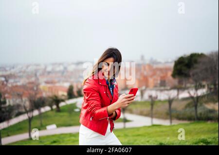 Fröhliche, lächelnde junge Frau, die ein modernes Smartphone benutzt und eine SMS oder textnachricht über ihr Mobiltelefon tippt, während sie an einem sonnigen Sommertag im Park spazieren geht. Stockfoto