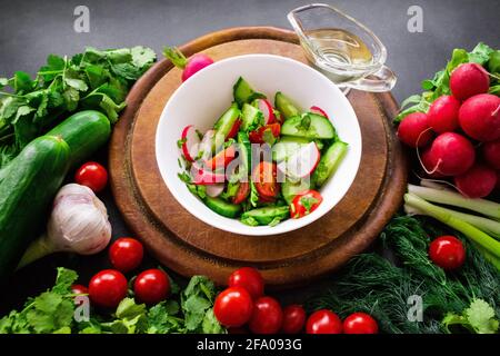 Ein Haufen frisches, reifes Gemüse: Radieschen, Tomaten, Gurken, Kräuter, Und Knoblauch sind um ein rundes Holzhackbrett angeordnet. Eine weiße Schüssel mit einem Witz Stockfoto