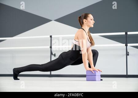 Seitenansicht einer atemberaubenden Tänzerin, die in der Tanzhalle Ausfallschritte übt. Wunderschöne Passform Frau trägt schwarze enge Kleidung Stretching Beine mit kleinen lila Blöcke auf dem Boden. Stretching, Gymnastik Konzept. Stockfoto