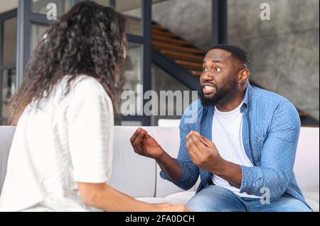 Irritierte schwarze Bursche, die die Frau anschreit, zorniger Freund streitet und gestikuliert mit verärgertem Gesichtsausdruck, afroamerikanische Paarstreitigkeiten sitzen zu Hause auf der Couch Stockfoto