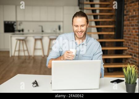 Fröhlicher Tausendjähriger arbeitet an Außenstunden, verwendet einen Laptop für den Empfang von E-Mails, führt Korrespondenz mit dem Kunden, entwickelt Software, die von zu Hause aus funktioniert, trinkt Kaffee und lächelt Stockfoto