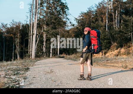 Eine Backpacker-Frau, die den Sonnenuntergang im Wald genießt. Eine Frau mit einem Rucksack, die sich umsieht und die Aussicht bewundert. Stockfoto