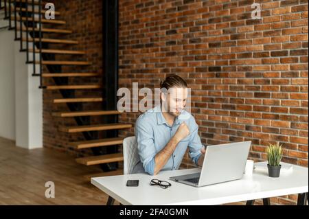 Ein lächelnder, junger Geschäftsmann blickt zufrieden auf den Laptop-Bildschirm, der am Schreibtisch im Loft-Bürobereich sitzt. Glad Freelancer erfand Startup-Idee Stockfoto