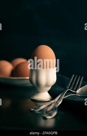 Einzelnes Ei in einem einzigen Eierbecher, eine Schüssel Eier im Hintergrund, dunkler Tisch und Hintergrund. Stockfoto