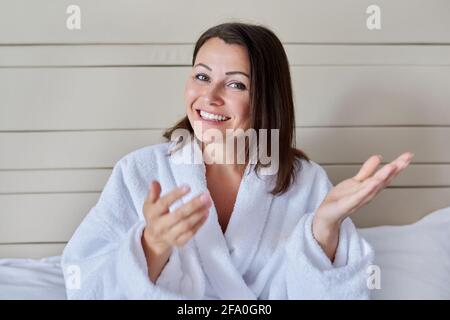 Eine freundliche Frau mittleren Alters in einem weißen Bademantel, die sich unterhielt und aussierte Bei der Kamera Stockfoto