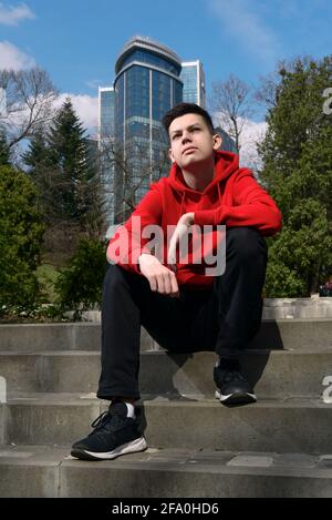 Ein gutaussehender, selbstbewusster Teenager, der am sonnigen Frühlingstag auf der Treppe im Stadtpark sitzt. Lässiger, jugendlich gekleideter roter Hoodie und schwarze Jeans Stockfoto