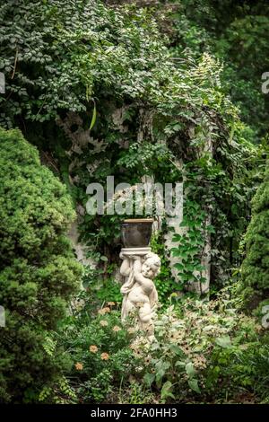 Amor Pflanzenstand hält Topfblume in Garten Nische mit Reben und mehr Blumen in der Nähe rustikalen Wand. Stockfoto