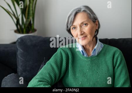 Ein Porträt einer charmanten strahlenden alten Dame, die posiert und die Kamera anschaut, grauhaarige ältere Frau, die im Wohnzimmer sitzt, einen lässigen grünen Pullover trägt, sich ausruht und lächelt Stockfoto