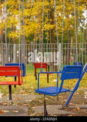 Leeres, altmodisches Karussell in einem Stadtpark. Rote und blaue Holzsitze auf Metallketten aus Schaukelkarussell. Bäume im Hintergrund, gefallene Blätter Stockfoto