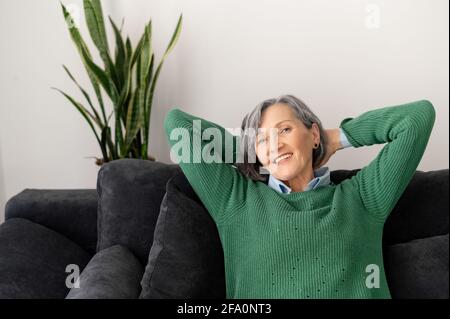Ein Porträt einer charmanten älteren Dame, die sich auf der Couch im Wohnzimmer entspannt. Eine Frau mittleren Alters, die sich hinter dem Kopf die Hände kreuzt, direkt auf die Kamera schaut und eine Pause von der Arbeit macht Stockfoto