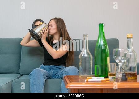 Junge dunkelhaarige Frau sitzt auf einem Sofa und trinkt nach einer stürmischen Party Wasser aus einer Teekane, leere Flaschen stehen neben ihr Stockfoto