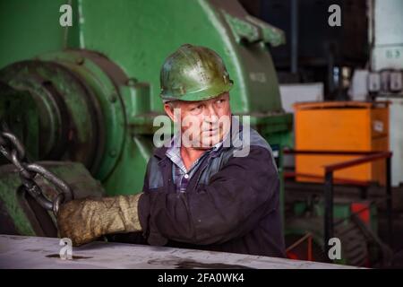 USt'-Kamenogorsk, Kasachstan:Fabrik Vostokmashzavod.Erwachsener kaukasischer Arbeiter mit grünem Hardhut.verschwommene grüne Maschine auf dem Hintergrund. Stockfoto