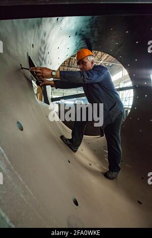 Ust-Kamenogorsk, Kasachstan. Fabrik Vostokmashzavod. Schwertechnikanlage. Arbeiter in einem riesigen Metallrohr. Orangefarbener Hardhut und graue Arbeitskleidung. Stockfoto