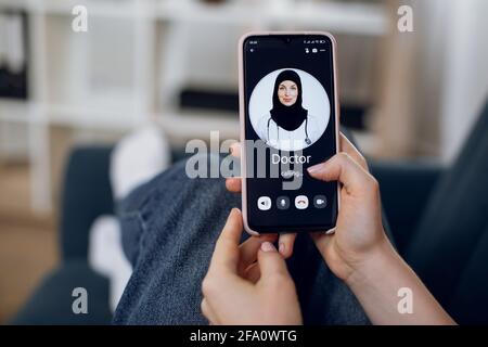 Nahaufnahme der Hand einer nicht erkennbaren weiblichen Patientin, das Mobiltelefon haltend und eine Videokonferenz mit ihrem arabisch-muslimischen Arzt in schwarzem Hijab. Online-Konsultation mit dem Arzt zu Hause. Stockfoto