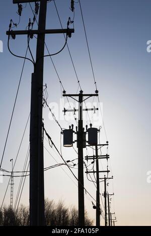 Reihe von verketteten Strommasten mit elektrischen Übertragungsleitungen und Transformatoren bei Sonnenaufgang Stockfoto