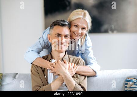 Familiäre Zärtlichkeit Beziehungen. Porträt eines älteren gutaussehenden kaukasischen Ehepaares in lässiger, stilvoller Kleidung, nette Frau, die im Wohnzimmer steht und ihren Mann umarmt, die Kamera anschaut und lächelt Stockfoto