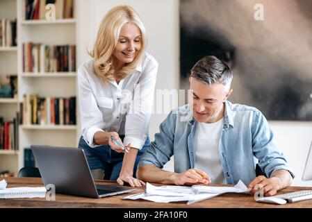 Zwei erfolgreiche, selbstbewusste, fröhliche kaukasische Geschäftspartner mittleren Alters in legerer Kleidung am Tisch, die Ideen und Strategien diskutierten, sich auf Dokumente konzentrierten und lächelten Stockfoto