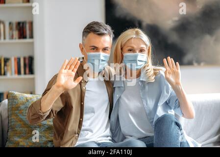 Gesundheitskonzept. Reifes kaukasisches Ehepaar, das Schutzmasken trägt, sich um die Gesundheit sorgt, auf dem Sofa sitzt, während der Videokonferenz mit Handgesten begrüßt und die Kamera anschaut Stockfoto