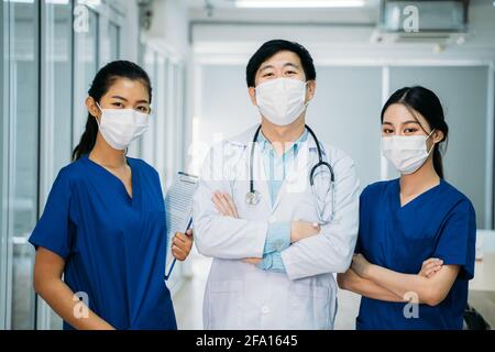 Porträt einer asiatischen Gruppe von Arzt und Krankenschwester In Uniform und Labcoat mit Stethoskop und Gesichtsmaske stehend Im Krankenhausgang mit gekreuzten Armen Stockfoto