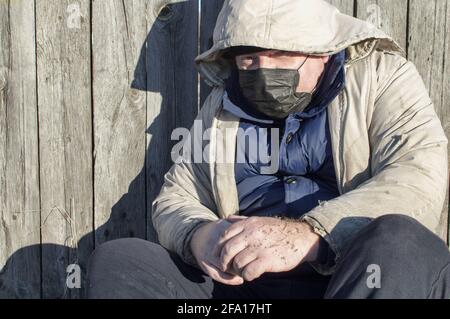 Obdachlos. Ein Mann in zerlumpter Kleidung und einer medizinischen Maske sitzt an einem Holzzaun. COVID-19. Nahaufnahme. Stockfoto