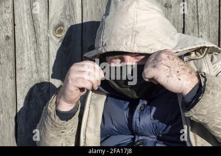 Obdachlos. Ein Mann in zerlumpter Kleidung und einer medizinischen Maske sitzt an einem Holzzaun. COVID-19. Nahaufnahme. Stockfoto