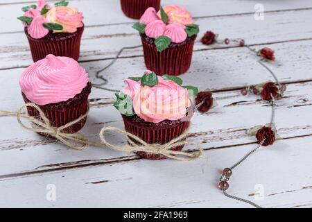 Nahaufnahme von roten Samt-Cupcakes, verziert mit Sisalfaden-Schleife, neben einer weiblichen Halskette mit kleinen Rosen. Stockfoto