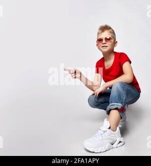 Frolic blonde Kid Boy in rotem T-Shirt, blauer Jeans, Turnschuhen und Sonnenbrille sitzt gehockt und zeigt mit dem Finger auf den Kopierraum Stockfoto