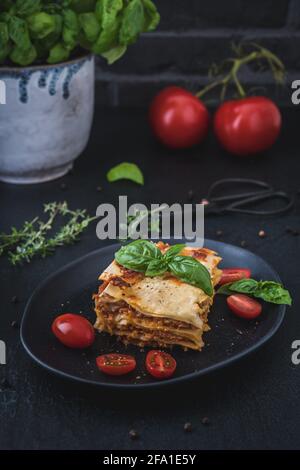 Stück selbstgemachte vegetarische Sojalasagne auf schwarzem Teller, Tomaten, Herben und Basilikum auf dunklem Hintergrund, vertikal Stockfoto