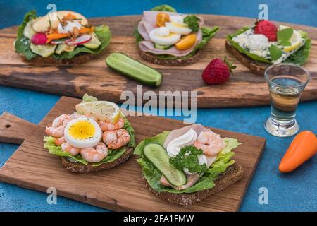 Verschiedene dänische offene Sandwiches auf Holzbrettern und blauem Hintergrund, dekoriert mit verschiedenen rohen Gemüse, Aquavit und Erdbeeren Stockfoto