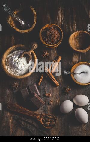 Verschiedene Backzutaten wie Mehl, Eier, Zucker und Kakao in Holzschüsseln auf einem dunklen Holztisch, vertikale Draufsicht Stockfoto