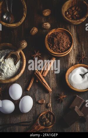 Vertikale Draufsicht auf verschiedene Backzutaten wie Mehl, Eier, Zucker und Kakao in Holzschüsseln auf einem dunklen Holztisch Stockfoto